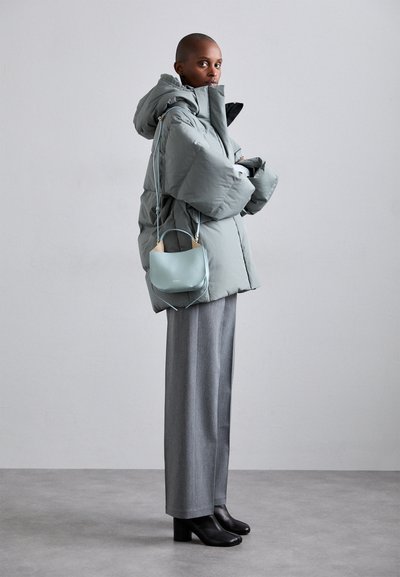 Doudoune gris clair avec un col montant, associée à un pantalon large gris et des bottes à talons noires. Un petit sac à main bleu clair pend à l'épaule.