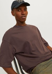 Brun kortærmet T-shirt med en afslappet pasform, parret med marmorgrønne denimbukser. En marineblå kasket med et emblem sidder på modellens hoved.