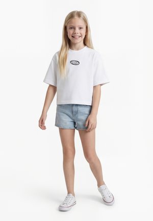 Smilende jente med langt blond hår, iført en hvit t-skjorte med svart logo, lyseblå denimshorts og hvite joggesko, stående på hvit bakgrunn.