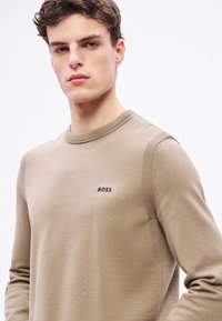 Beżowy sweter z długim rękawem, z prążkowanym, okrągłym dekoltem, z subtelnym logo „BOSS” na piersi oraz teksturowanym splotem na ramionach.