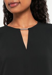 Woman wearing a black top with a small V-neck slit held by a horizontal silver bar detail at the neckline.
