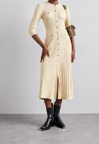 Virkat, krämfärgat klänning med trekvartslånga ärmar, knappfront och texturerat mönster; matchas med svarta ankelskorte stövlar och en liten handväska.