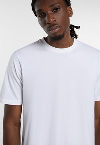 Young man with dark skin and dreadlocks wearing a plain white short-sleeve t-shirt against a white background.