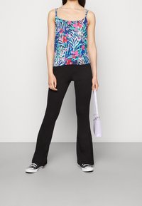 Top con fiori multicolori e spalline regolabili, abbinato a pantaloni neri a zampa d'elefante e sneaker nere; tiene una borsa viola chiaro.