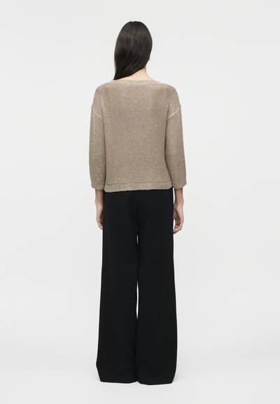 Femme aux longs cheveux foncés portant un pull beige tricoté à manches trois-quarts et un pantalon noir à jambes larges, debout de dos.