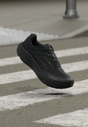 Zapatilla de correr negra con suela gruesa, flotando sobre un paso de peatones en una calle con pequeñas piedras volando alrededor.