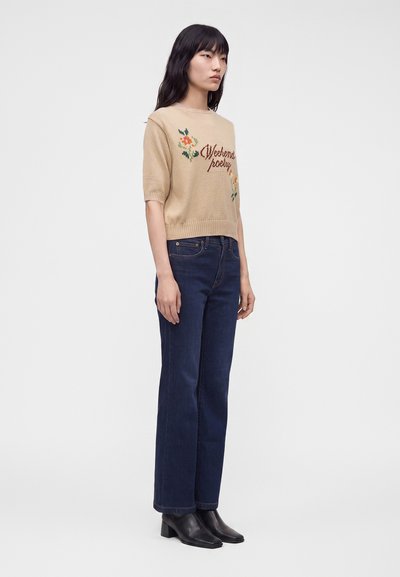 Femme aux longs cheveux noirs portant un pull beige à manches courtes avec un motif floral, un jean bleu foncé et des bottines noires, debout de profil.