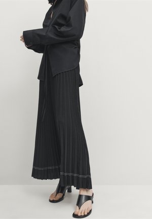 Woman wearing black pleated wide-leg pants, oversized black shirt, and black heeled sandals with toe straps against a plain background.