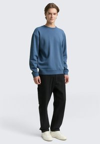 Niebieski sweter z okrągłym dekoltem o luźnym kroju, z ściągaczami przy mankietach i u dołu. Noszony z czarnymi spodniami w luźnym kroju i białymi trampkami. Gładki wzór, miękka tekstura.