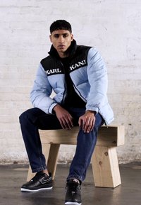 Lyseblå dunjakke med svarte detaljer og "KARL"-tekst, svarte jeans og svarte joggesko, sittende på en trebenk mot en hvit vegg.