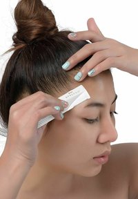 Vitt stylingverktyg som används för att forma ögonbryn, hålls mellan fingrarna med ljusblå nagellack. Håret är uppsatt i en knut mot en enfärgad bakgrund.