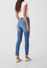 Jeans de denim azul ajustados con una cintura alta, color desvaído cerca de los tobillos y dobladillos deshilachados, combinados con una camiseta blanca ajustada y zapatos claros.