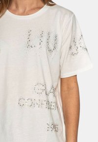 T-shirt bianco a maniche corte con applicazioni di strass che formano schemi di testo. Tessuto morbido con vestibilità rilassata e scollatura rotonda.