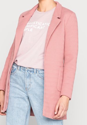 Person trägt einen strukturierten pinkfarbenen Blazer, ein hellrosa Hemd mit weißem Text sowie hellblaue, hoch taillierte Jeans mit Taschen und Gürtelschlaufen.