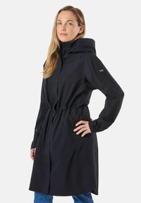 Žena oblečená v čiernom dlhom pršiplášti s kapucňou, s obtiahnutým pásom a vreckami na zips, v kombinácii s modrými džínami, stojaca pred bielym pozadím.