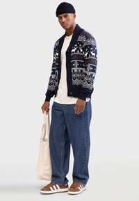 Cardigan tricotat bleumarin cu modele albe de iarnă, tricou bej, blugi largi albaștri, pantofi maronii cu dungi albe și o geantă din pânză.