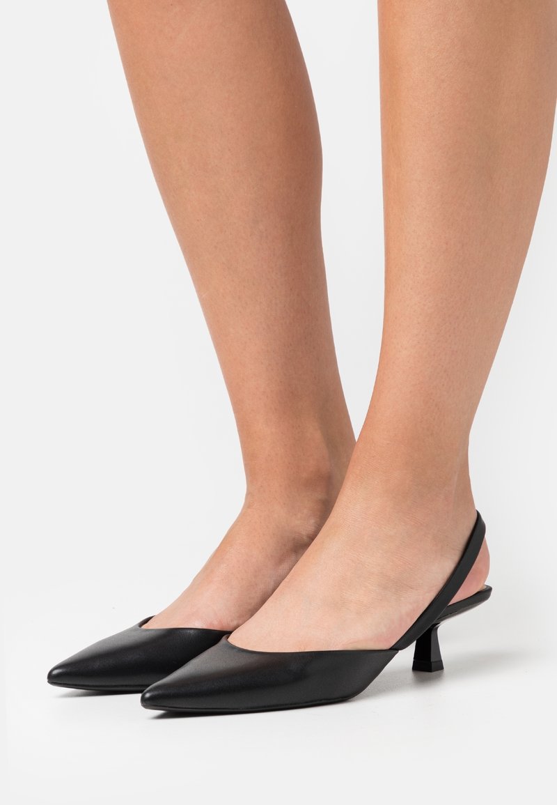 Personne portant des escarpins slingback noirs à bout pointu avec des talons carrés bas sur une surface blanche.