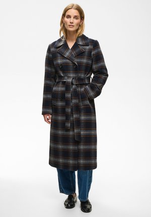 Femme portant un long manteau ceinturé à carreaux foncé par-dessus un jean bleu et des mocassins noirs, debout avec une main dans la poche devant un fond blanc.