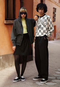 Haut de survêtement gris surdimensionné, chemise à manches longues noire et blanche à motifs, jupe plissée noire, collants noirs et baskets noires avec semelles blanches.