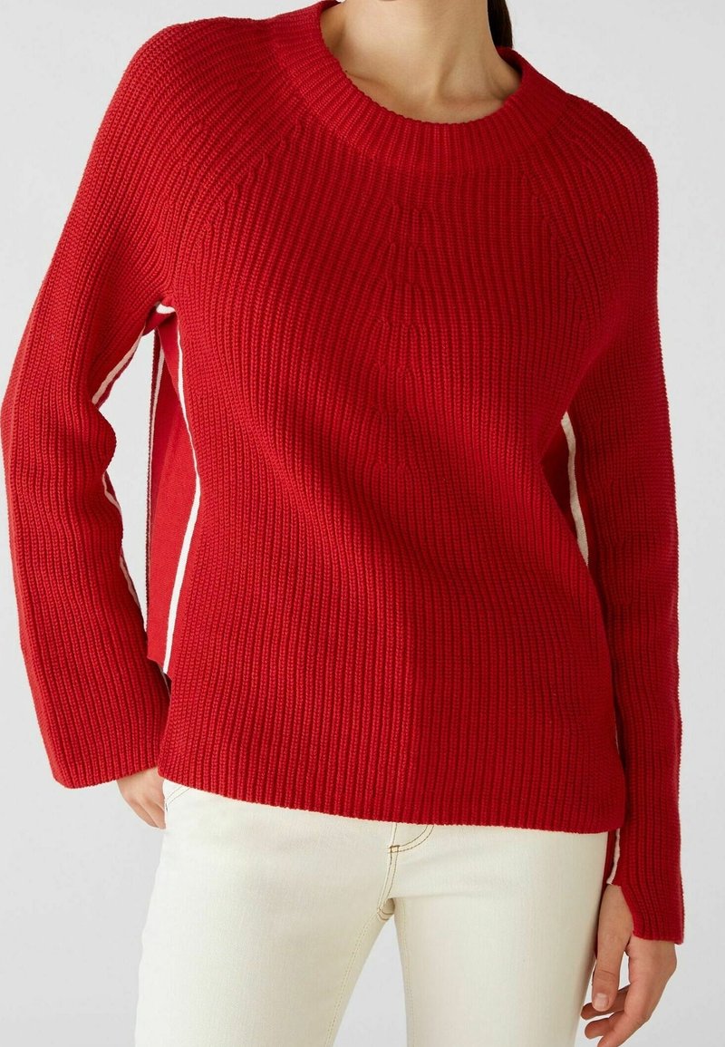 Gros plan d'une personne portant un pull en maille côtelée rouge épais à manches longues et un pantalon blanc sur un fond uni.