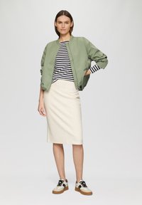 Veste bomber verte portée sur une chemise rayée, associée à une jupe midi crème et des baskets avec des accents beige, noir et blanc.