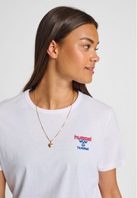 Ung kvinde med langt mørkt hår iført en hvid Hummel t-shirt og gyldt hjerteformet vedhæng, kigger mod højre mod en ensfarvet baggrund.