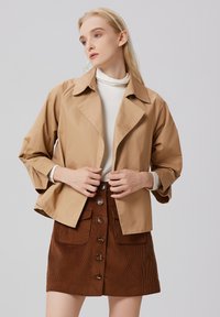Veste courte beige à col cranté, associée à une jupe en velours côtelé marron avec des boutons sur le devant et des poches.