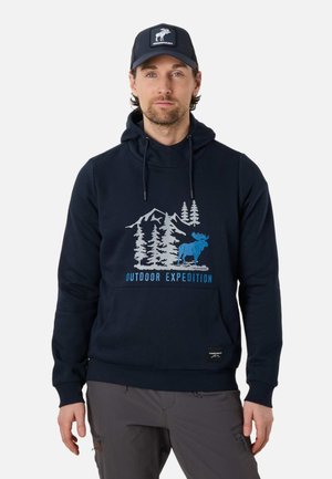 Mand iført marineblå hættetrøje med grafik af bjerg, træer og elg, teksten "Outdoor Expedition" og matchende marineblå kasket med elg-logo.