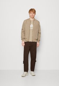 Beige Bomberjacke mit gerippten Bündchen und Saum, über einem hellbraunen T-Shirt mit einem weißen Logo. Dunkelbraune Hose mit seitlichen Akzenten. Weiße Sneakers.