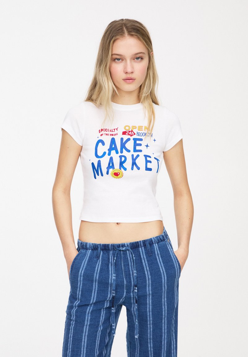 Hvid cropped t-shirt med farverig tekst, der står "CAKE MARKET", parret med blå stribede bukser. Fremstillet af bomuld, kortærmet, afslappet pasform.