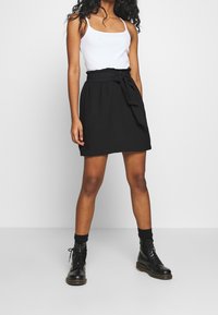 Jupe portefeuille noire avec un détail de taille nouée, associée à un débardeur blanc. Le mannequin porte des bottines noires à lacets et des chaussettes foncées.