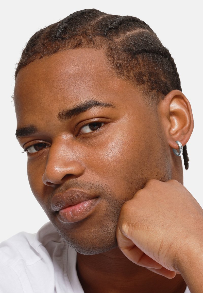 Nærbilde av en person med flettet hår, iført en sølvringproppe. Huden er glatt med naturlige nyanser; skjorten er hvit. Hånden hviler på haken.