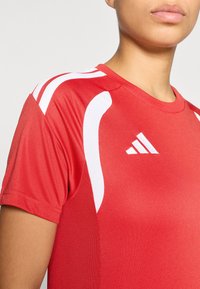 Κόκκινο αθλητικό πουκάμισο με λευκές λεπτομέρειες και διακριτικό διάτρητο ύφασμα στα μανίκια, με στρογγυλή λαιμόκοψη και το λογότυπο της Adidas στο αριστερό στήθος.