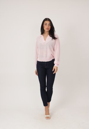 Donna con capelli scuri che indossa una maglia a maniche lunghe rosa chiaro, jeans skinny scuri e tacchi bianchi aperti in punta, in piedi davanti a uno sfondo semplice.