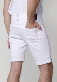 Pantalones cortos de denim blanco con un dobladillo enrollado y detalles en costura roja, que cuentan con un bolsillo trasero y un bolsillo lateral para la mano. Textura suave.