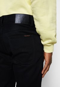 Schwarze Jeans aus Denim mit strukturiertem Finish, ausgestattet mit einem schwarzen Lederetikett am Bund und orangen Nähten an der Gesäßtasche. Gelbes Sweatshirt.