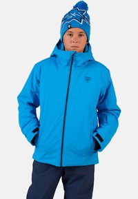 Modrá voděodolná bunda s kapucí, zipovým zapínáním a elastickými manžetami. Nosit s námořnickými kalhotami. Čepice má hvězdicový vzor a bambuli.