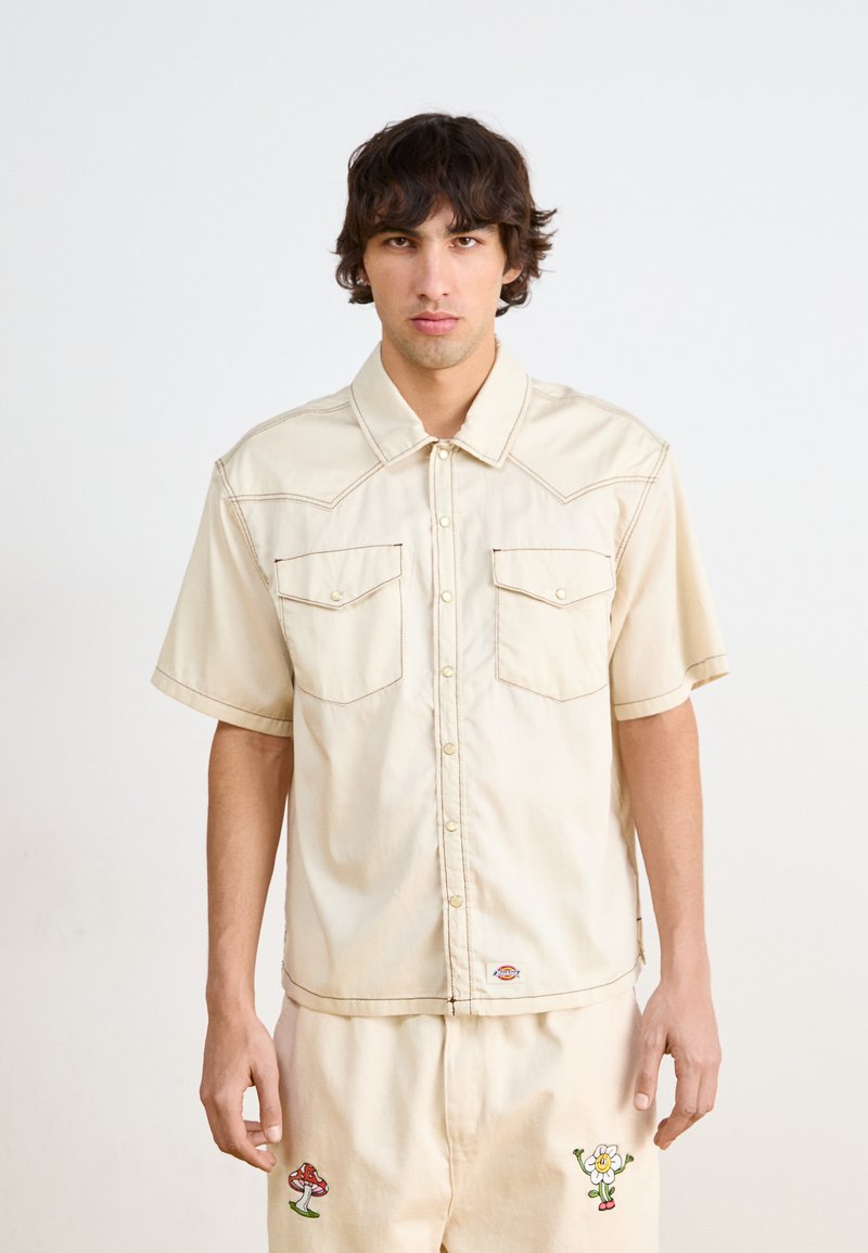 Camicetta beige a maniche corte con due tasche frontali, cuciture a contrasto e colletto a punta. Indossata con pantaloni beige abbinati con disegni ricamati.