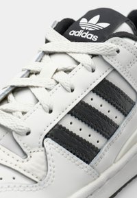 Baskets blanches avec accents noirs, dotées de trois bandes horizontales, cuir texturé, fermeture à lacets et une languette noire avec logo.