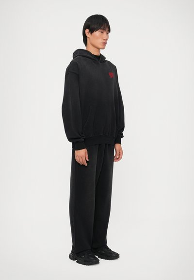 Sudadera negra con capucha, bolsillo frontal y logo rojo; combinada con pantalones anchos negros y zapatillas negras sobre un fondo liso.