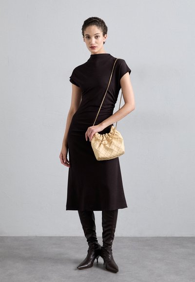 Rochie neagră, ajustată, cu mâneci scurte, asortată cu cizme maro până la genunchi și o geantă bej matlasată. Modelul stă în fața unui fundal gri.