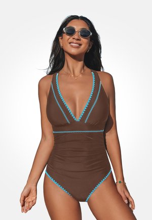 Donna con costume intero marrone con bordi blu, occhiali da sole e gioielli, sorridente su uno sfondo bianco uniforme.