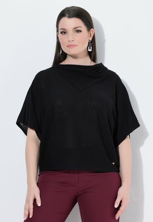 Frau mit langen dunklen Haaren, die ein schwarzes kurzärmeliges Oberteil und bordeauxrote Hose trägt, steht vor einem einfarbigen hellen Hintergrund.