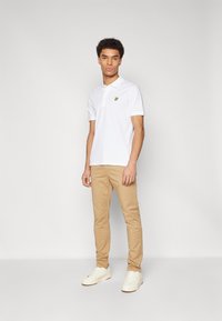 Hvit polo med en liten gul logo, kombinert med beige slim-fit bukser og hvite joggesko på en nøytral bakgrunn.