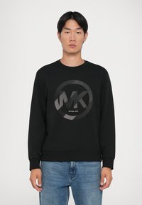 Μαύρη μπλούζα με στρογγυλή γκρίζα γραφική απεικόνιση που διαθέτει το κείμενο "MK" και "MICHAEL KORS". Ριμπ τελείωμα στον λαιμό και στα μανίκια, φτιαγμέν�η από λείο ύφασμα.