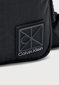 Μαύρη τσάντα χιαστί Calvin Klein με υφή υφάσματος, γεωμετρικό μοτίβο και λαστιχένιο σήμα με ασημένια γράμματα.