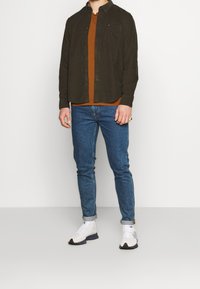 Mörkgrön skjorta med knappar över en rostfärgad t-shirt, kombinerad med blå smala jeans och vita träningsskor.