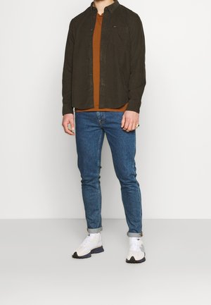 Mörkgrön skjorta med knappar över en rostfärgad t-shirt, kombinerad med blå smala jeans och vita träningsskor.