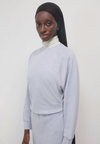 Eine Frau trägt ein schwarzes Kopftuch, einen weißen Rollkragenpullover und ein hellgraues Sweatshirt und steht vor einem schlichten weißen Hintergrund.