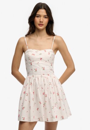 Jeune femme portant une robe blanche sans manches avec de fines bretelles et un motif floral rouge mêlé de pois, debout les mains derrière le dos.